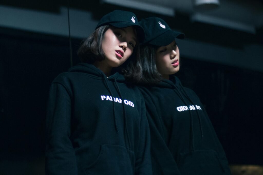 A fashionable woman poses with her reflection in a dark, moody setting wearing a hoodie and cap.